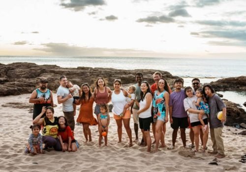 A large multiracial group of adults and children stand on a sandy beach in Hawaii at sunset and smile directly at the camera.